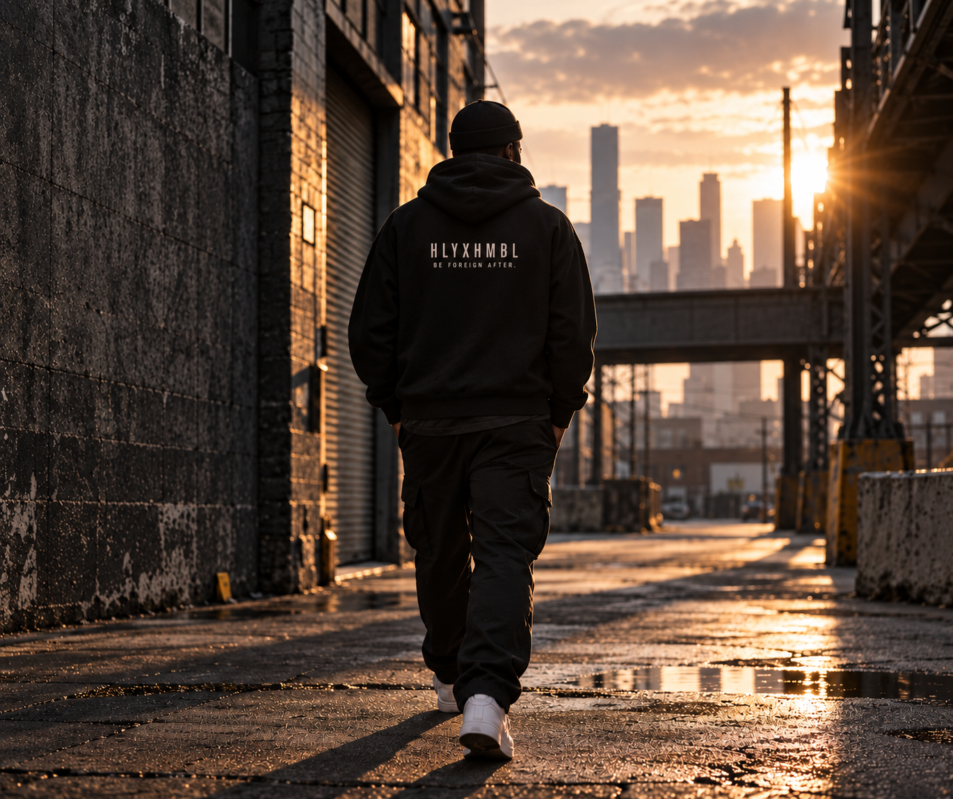 Model walking through a sunlit alley in a black HLYXHMBL hoodie